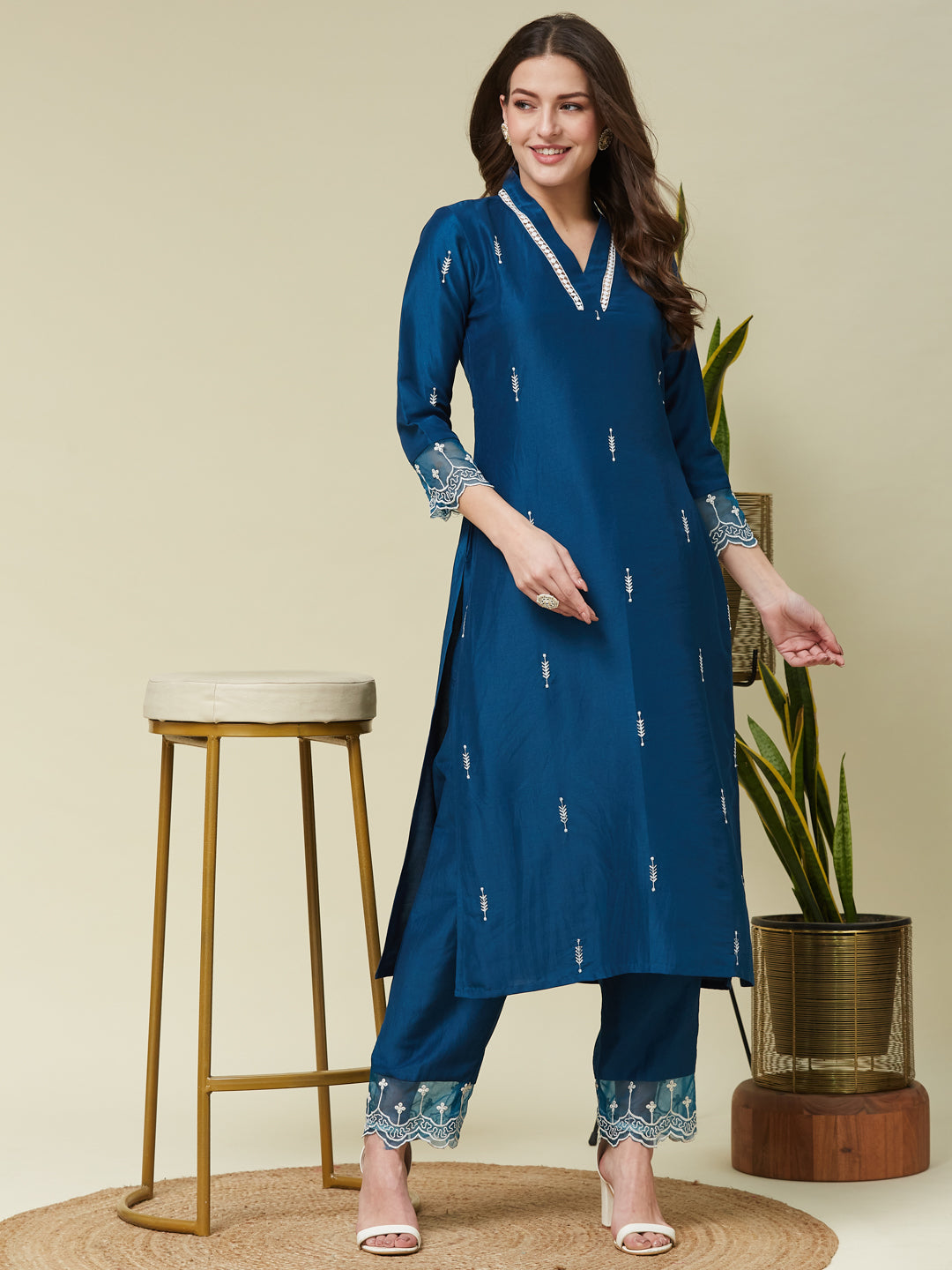 A woman modeling a teal blue chinon embroidered co-ord set with delicate floral patterns and sequins, standing beside a stool and a potted plant. Teal Blue Chinon Embroidered Co-ord Set