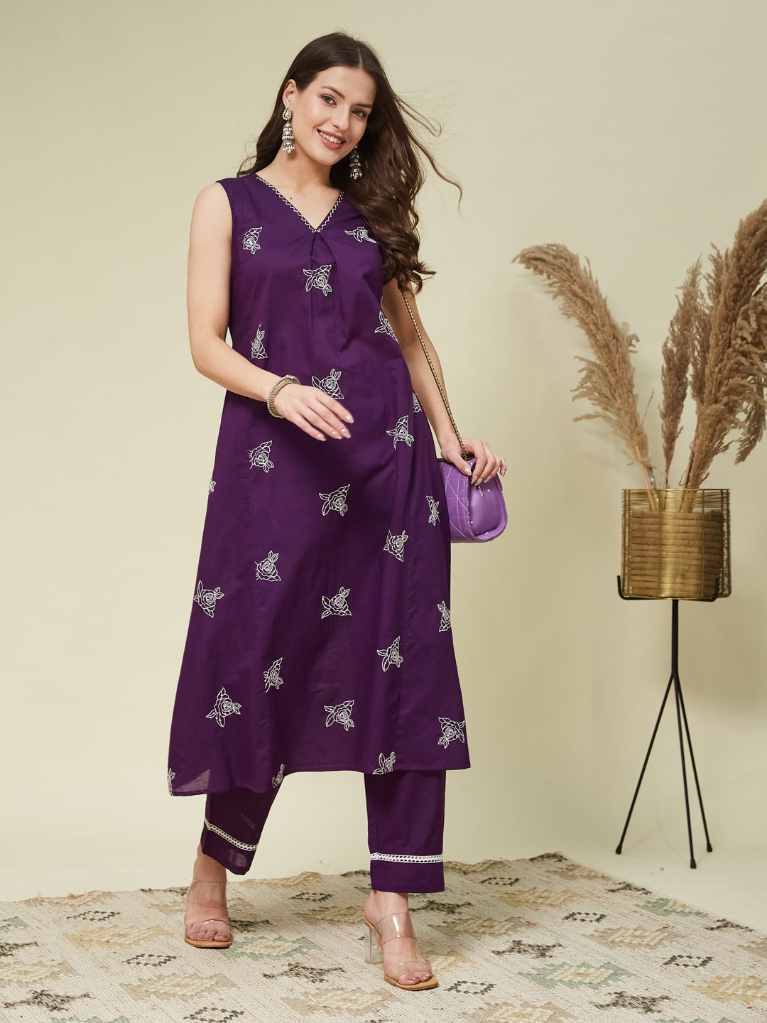 A woman wearing a sleeveless purple top with floral embroidery and matching pants, standing in a room with a neutral background.