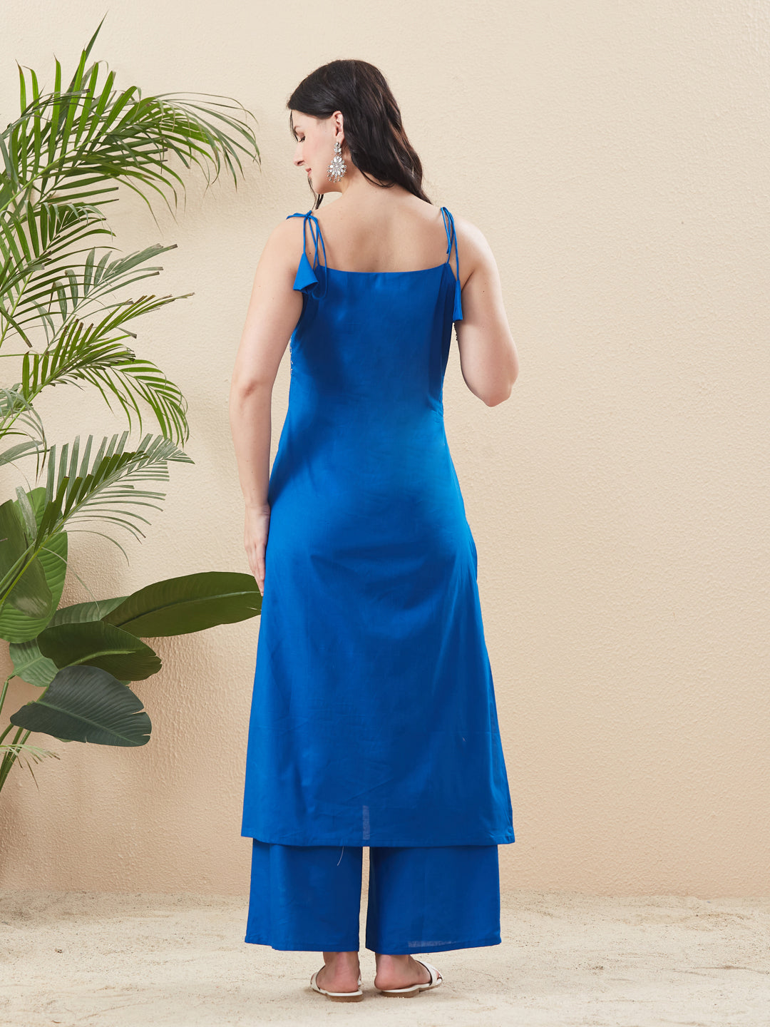 Woman wearing a blue dress standing against a beige wall with a plant to the left. Blue Cotton Embroidered Kurta with Palazzo Set for Women | backside view