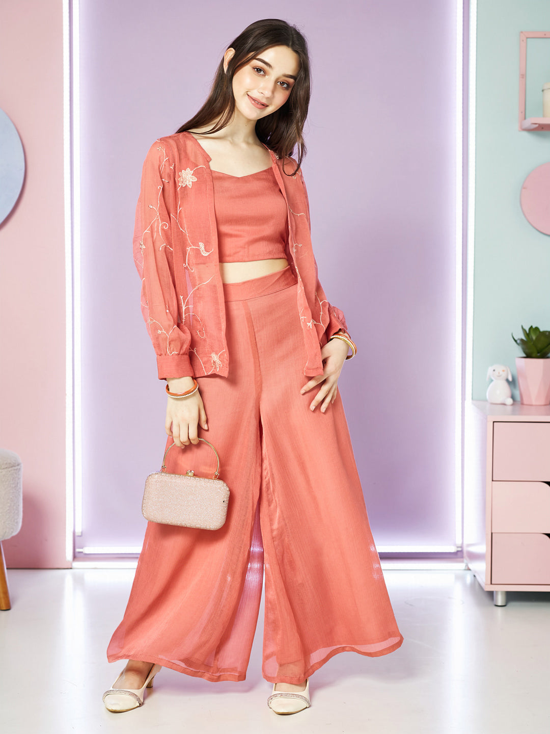 Woman in a coral outfit with a matching handbag in a pastel- Women Pink Crop Top with Palazzo and Shrug Setcolored room.